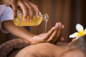 Close-up of golden massage oil being poured into a hand in a spa setting, with a person lying on a massage table and a plumeria flower near their ear.