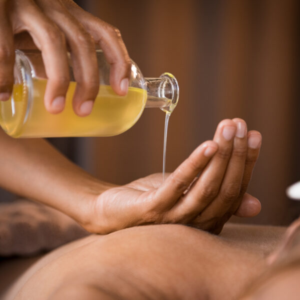 Close-up of golden massage oil being poured into a hand in a spa setting, with a person lying on a massage table and a plumeria flower near their ear.